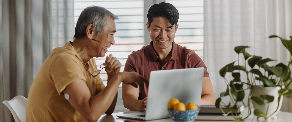 A man wearing a FreeStyle Libre sensor on the upper arm while using a laptop with another man at a table.