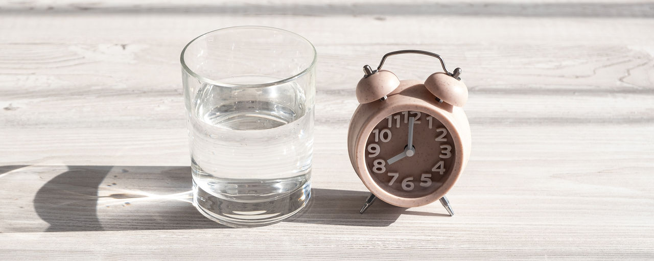alarm clock and sleep mask, glass with clean water on a wooden white table in the morning sunlight. The concept of starting a new day, good morning or ending the day, late evening. Day planning. High quality photo