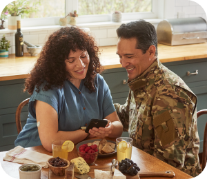 A woman, wearing a FreeStyle Libre sensor, and a man, dressed in a military uniform, sit together at a kitchen table smiling.