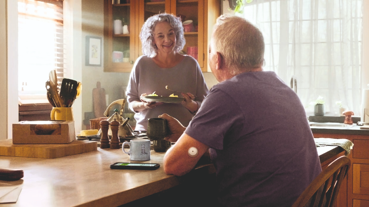 Κύριος με αισθητήρα FreeStyle Libre 2 Plus πίνει καφέ, έχοντας το κινητό με την εφαρμογή FreeStyle LibreLink στο τραπέζι δίπλα του, και συζητάει με τη σύζυγό του η οποία χαμογελάει