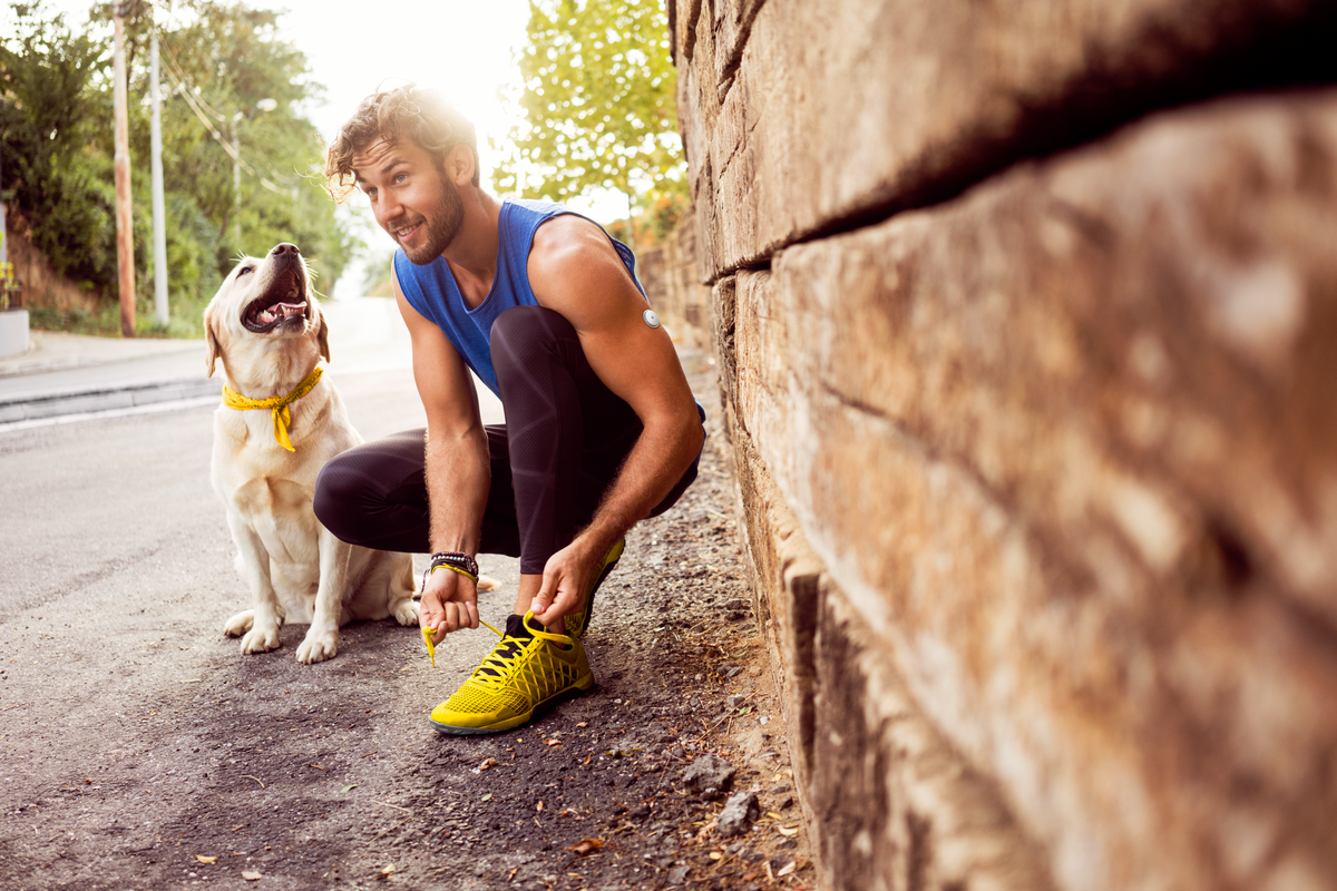 Νεαρός άντρας με αισθητήρα FreeStyle Libre 2 Plus που αθλείται παρέα με το σκύλο του, δένει τα κορδόνια του παπουτσιού του