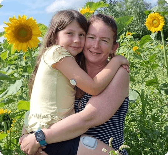 A mother holding her daughter in front of a sunflower field. The daughter's FreeStyle Libre sensor is visible on the back of her upper arm.