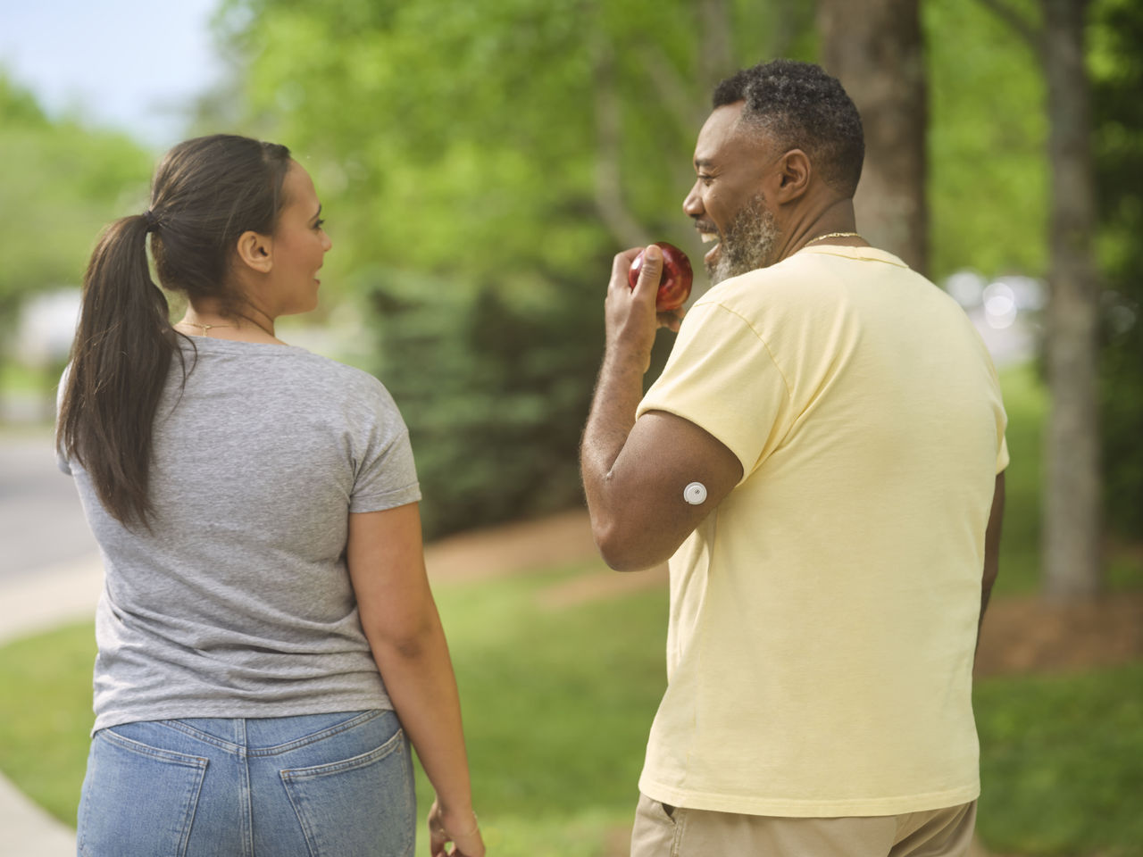 Un homme avec un capteur sur le haut du bras mangeant une pomme et se promenant dans le parc avec son ami.