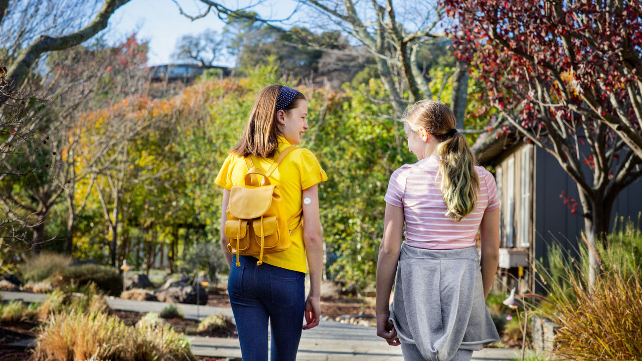 two girls walking outside, smiling and laughing at each other. The girl wearing the yellow shirt is wearing FreeStyle Libre 3 sensor on her arm.