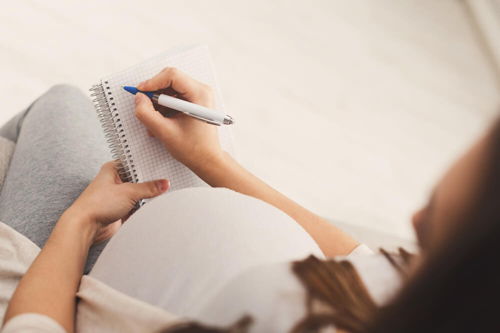 Unrecognizable pregnant woman lying on sofa with blank notebook and pen and writing names for her baby, making shopping list, top view, copy space