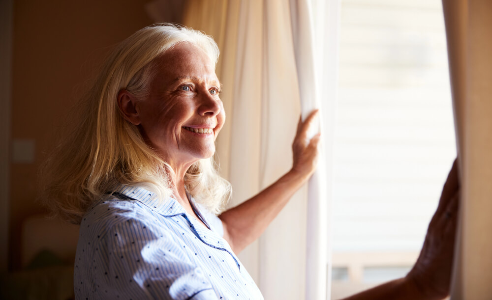 Smiling senior white woman opening the curtains on a sunny morning, side view, close up