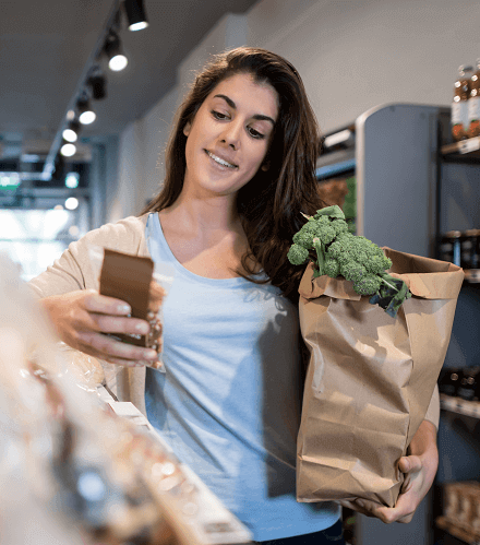 Kobieta w sklepie spożywczym w jednym ręku trzyma papierową torbę z produktami spożywczymi, uśmiecha się. W drugim ręku trzyma telefon z aplikacją LibreLinkUp.