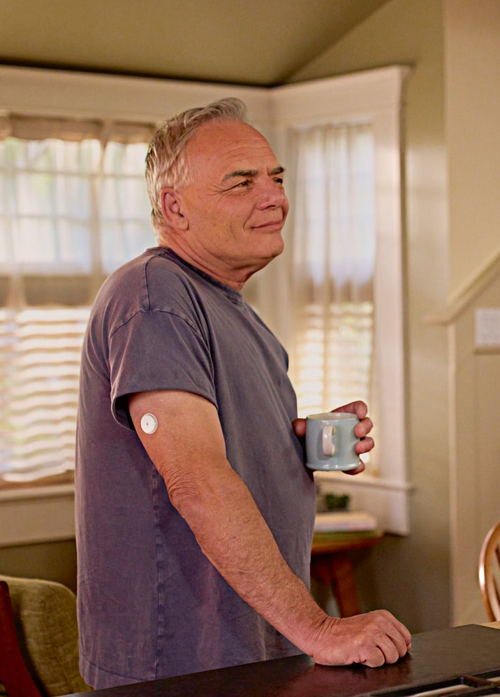 Nastolatek z sensorem FreeStyle Libre 2 na ramieniu siedzi przy okrągłym stole naprzeciwko kobiety w żółtej koszuli, która uśmiecha się do niego i gestykuluje. Na stole stoją szklanki z wodą, a w tle widoczny jest plakat pokazujący anatomię człowieka.