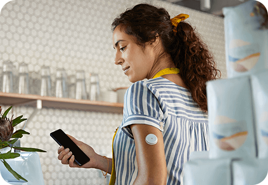 Kvinna står i ett kök med sensor på armen och kollar på sin telefon