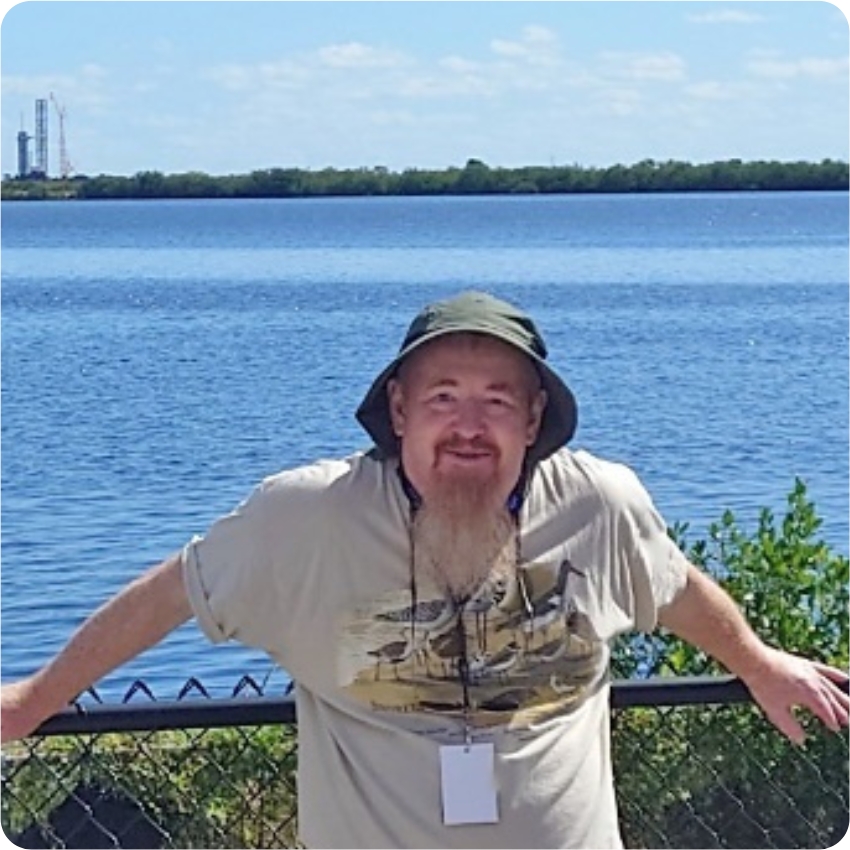 Mark Tiller wearing a t-shirt and hat, stood in front of a body of water, leaning against a fence
