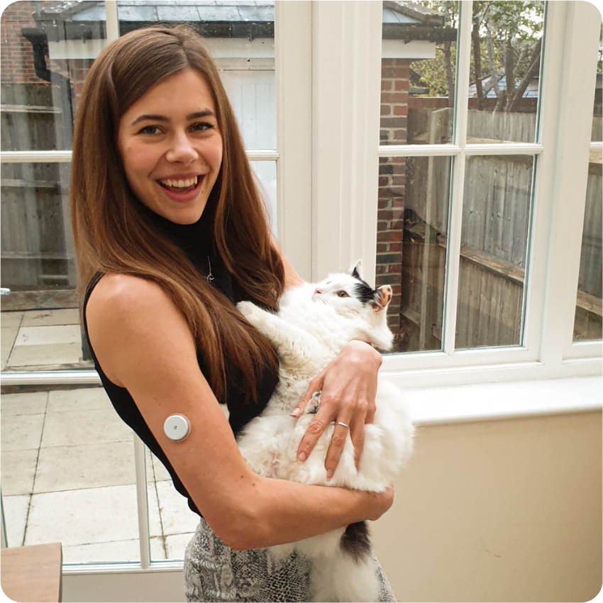 Elise Quarrington standing in a conservatory holding her black and white cat and FreeStyle Libre sensor visible on the back of her upper arm