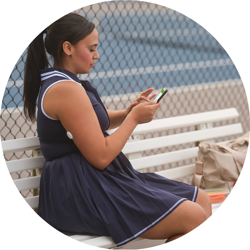 Woman in a navy tennis dress sitting on a bench beside a court, checking her phone. A FreeStyle Libre 2 Plus glucose sensor is visible on her upper arm.