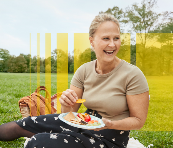 A woman sitting downs on a picnic blanket and holding a plate of food and smiling. Her FreeStyle Libre sensor is visible on the back of her arm.