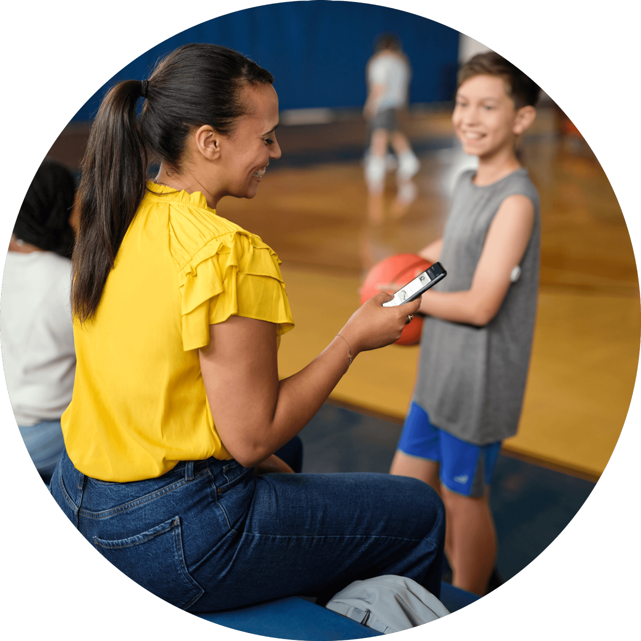 Mother checking LibreLinkUp app while her son with a FreeStyle Libre sensor holds a basketball in a gym